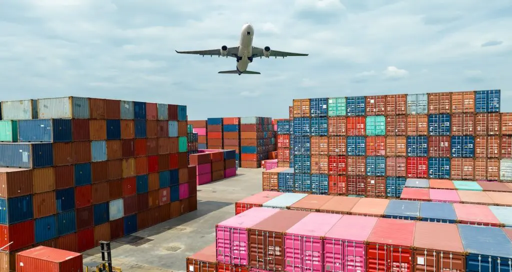 An aeroplane with air freight on board flying over a seaport with sea freight. These are examples of how freight forwarders ship.